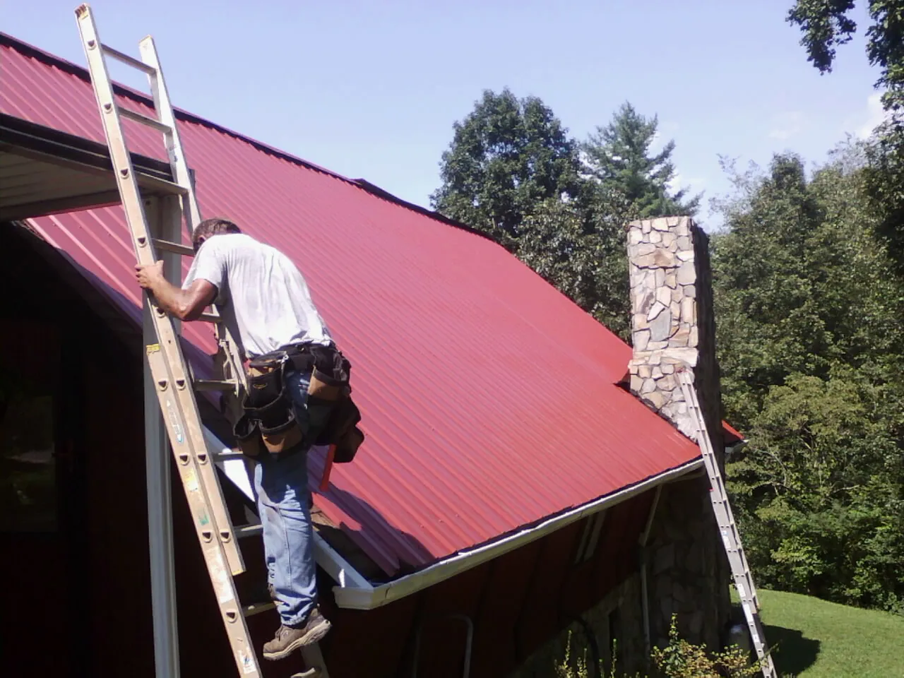 Roofer on a ladder beside a red metal roof and stone chimney — Soffit & Fascia Repair by Roofing Brothers in Mcpherson
